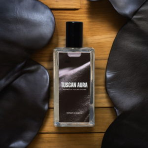 A Perfume named "Tuscan Aura" is lying on a Wodden Table with the surroundings of Black Tuscan Leather Jackets